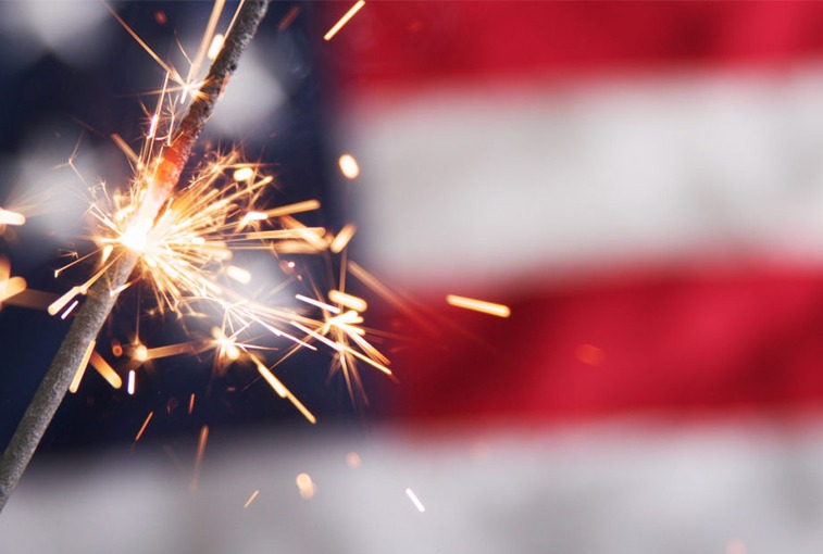 Someone is celebrating Independence Day, and that's why he is holding a burning sparkler. On background, we can see the flag of the United States of America.