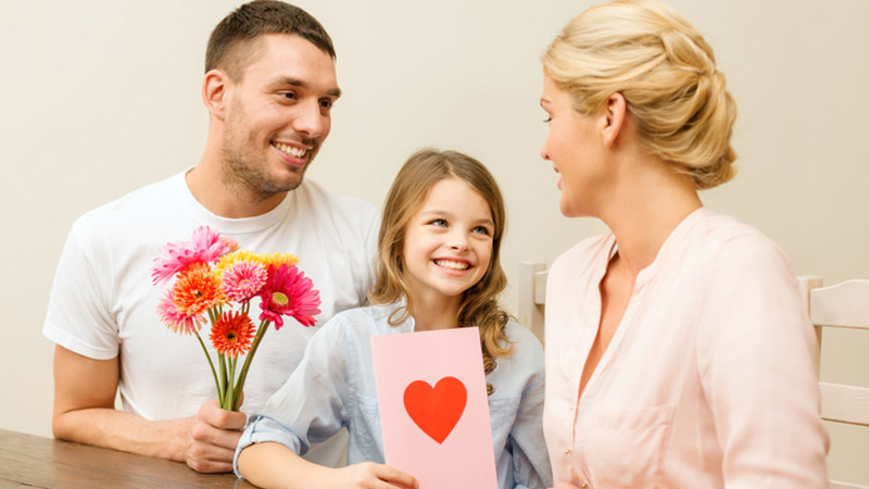 A family of three celebrates Mother's Day. Father is holding the flowers, and the daughter shows a postcard and the mother is happy to be in the center of attention.