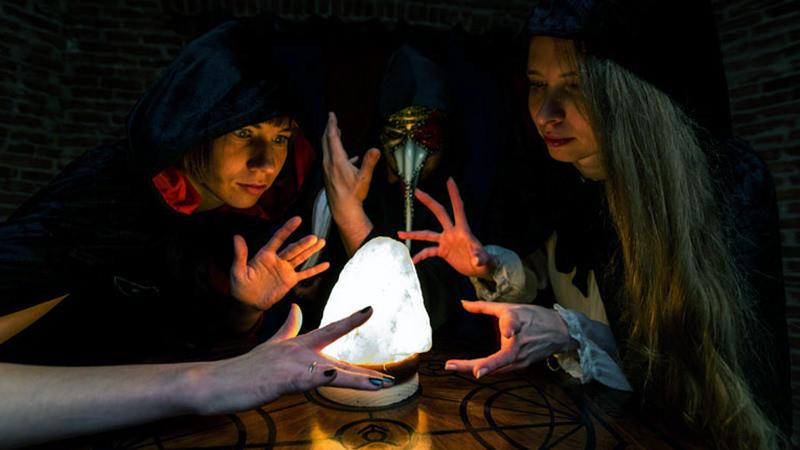 Two young ladies try to figure out the right solution to the magical puzzle in a mystical escape room. They are dressed in black robes, so they need to think like witches to progress further.